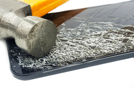 A close-up image of a hammer smashing a smartphone, resulting in a severely cracked and shattered screen. The photo highlights the concept of device destruction, damage, and the fragility of modern technology.の写真素材