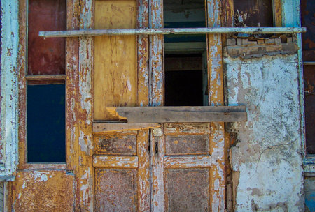 Close up of a crumbling and boarded up wooden door with cracked, peeling paint in shades of yellow, blue and brown.の写真素材