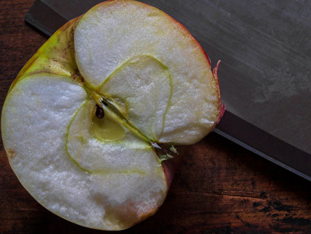 Close-up of a halved apple on a rustic wooden surface, with a large knife beside it. The image highlights the fruit's juicy, crisp interior and seeds.の写真素材