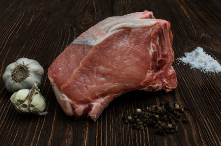 An image of a raw pork chop placed on a rustic dark wood surface, accompanied by garlic, a sprinkle of salt and a mixture of whole peppersの写真素材