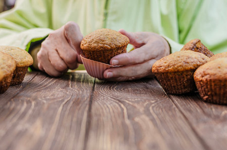 A close-up view of a hand holding a freshly baked muffin over a rustic wooden table, highlighting the texture and warmth of the pastry.の写真素材