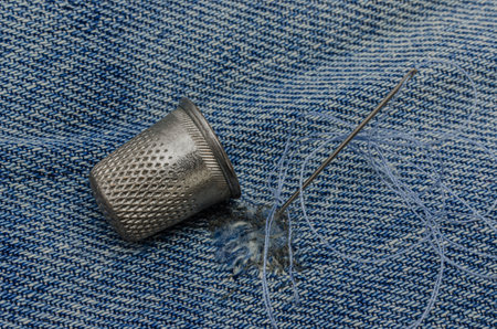 Close-up image of a metal thimble, sewing needle with thread, and a denim fabric showing visible stitching work, symbolizing traditional textile repair or hand sewing techniques.の写真素材