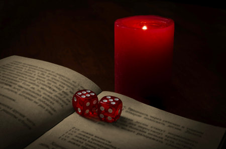 A dark, atmospheric composition featuring two red translucent dice placed on an open book illuminated by the warm glow of a red candle. The soft lighting and shadows create a moody, mystical ambiance, suggesting themes of chance, fate, or ritual.の写真素材