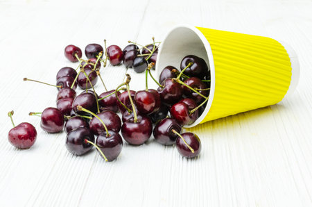 Ripe, glossy cherries spill out of a bright yellow paper cup onto a white wooden surface.の写真素材