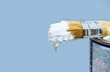 A close-up shot of a paintbrush with white paint dripping from its bristles, resting on the edge of a paint can against a clean, light blue background.の写真素材