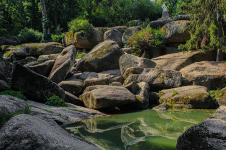 A tranquil green pond reflects the surrounding lush trees and ancient moss-covered boulders, creating a peaceful and picturesque natural landscape.の写真素材