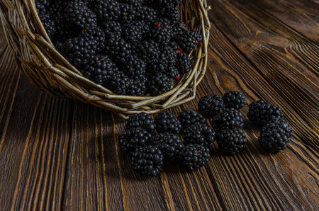 A close-up shot of ripe, dark blackberries spilling out of a rustic woven basket onto a rich, textured wooden surface, highlighting the freshness and abundance of the summer harvest.の写真素材