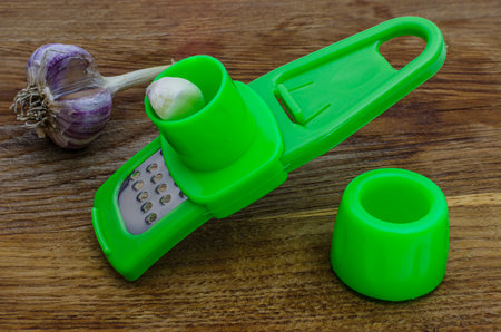 A close-up shot of a green plastic garlic grater with a garlic clove inserted, next to a whole head of garlic on a wooden background.の写真素材