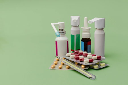 Close-up of various medical supplies including nasal sprays, throat spray, blister packs of pills, capsules, and a mercury thermometer on a green background, representing healthcare and treatment.の写真素材