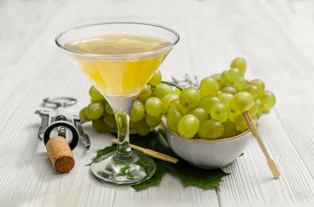 A bright, clear glass of white wine stands next to a small white bowl filled with fresh green grapes, against a rustic white wood backdrop. A cork and corkscrew are visible to the left.の写真素材
