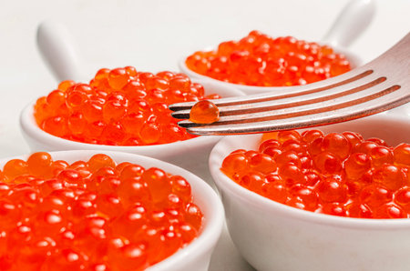Close-up of red salmon caviar in white bowls with a fork holding one roe grain. Gourmet seafood...