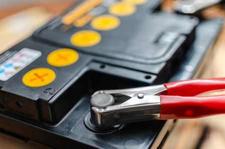 Close-up of a 12v automotive lead-acid battery being charged. A red metal crocodile clamp is connected to the positive terminal of the black battery casing with yellow caps.の写真素材