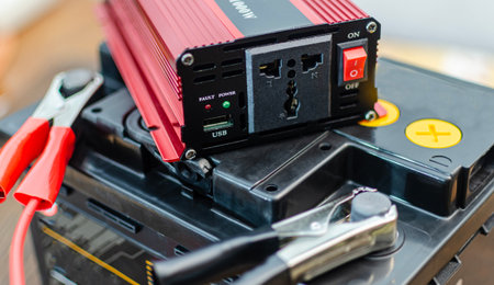 A close-up shot of a red and black power inverter connected to a car battery using heavy-duty jumper cables with alligator clips. The inverter features standard AC power outlets, a USB charging port, and status indicator lights.の写真素材