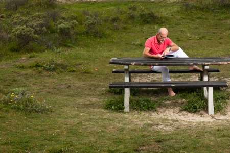 Retired senior man resting and using his tablet at table in park outdoors in grass dune landscape.の写真素材