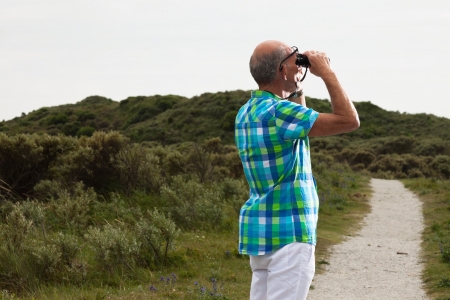 Senior man with beard and glasses using binoculars outdoors in grass dune landscape. Wearing green blocked shirt.の写真素材
