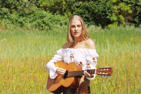 Retro blonde 70s hippie girl with acoustic guitar outdoor in nature. Standing in meadow.の写真素材