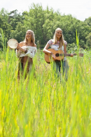 Two retro blonde 70s hippie girls making music with acoustic guitar and tambourine outdoor in nature.の写真素材