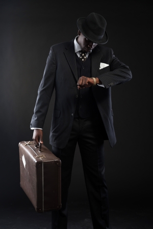 Retro african american traveller wearing striped suit and tie and black hat. Holding suitcase and looking on his watch. Studio shot.の写真素材