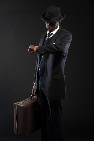 Retro african american traveller wearing striped suit and tie and black hat. Holding suitcase and looking on his watch. Studio shot.の写真素材