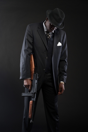 Retro african american mafia man wearing striped suit and tie and black hat. Holding machine gun. Studio shot.の写真素材