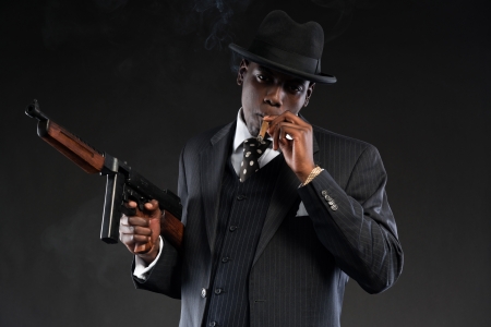 Retro african american mafia man wearing striped suit and tie and black hat. Smoking cigar. Holding a gun. Studio shot.の写真素材
