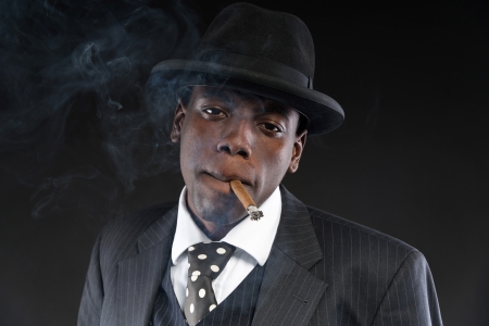 Retro african american mafia man wearing striped suit and tie and black hat. Smoking cigar. Studio shot.の写真素材