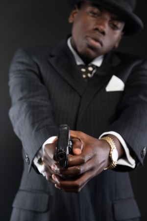 Retro african american mafia man wearing striped suit and tie and black hat. Shooting a
with gun. Studio shot.の写真素材