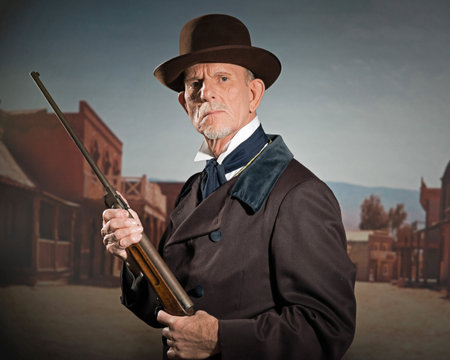 Senior western man wearing a brown hat and coat holding rifle. Standing in old small cowboy town.