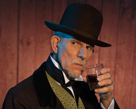 Senior alert western man wearing a brown hat and coat holding a whiskey. In front of wooden wall in...