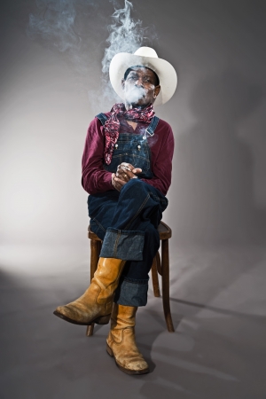 Retro senior afro american blues man in times of slavery. Wearing denim bib and brace overall with white hat. Smoking a cigarette.の写真素材