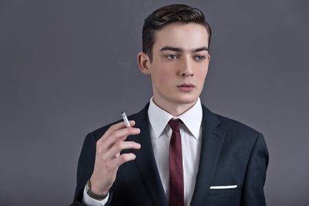 Retro fashion fifties young businessman wearing dark suit and tie. Smoking a cigarette. Studio shot against grey.の写真素材