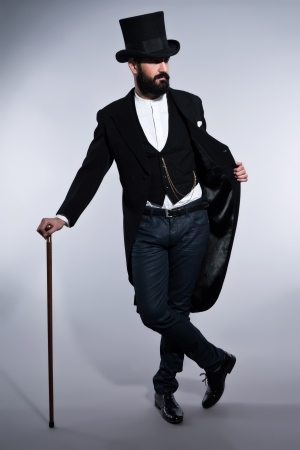 Retro hipster 1900 fashion man in suit with black hair and beard. Wearing black hat. Standing with cane. Studio shot against grey.の写真素材