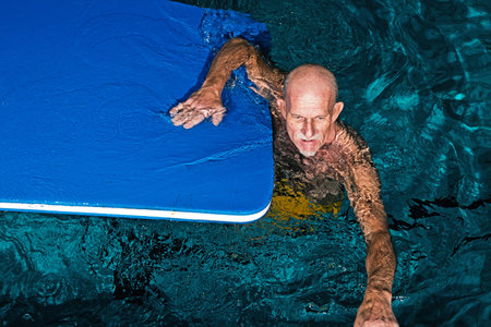 Healthy active senior man with beard in indoor swimming pool playing with foam raft.の写真素材