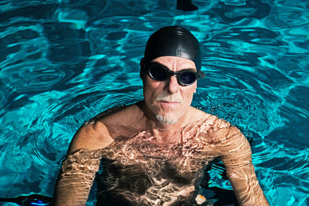 Active healthy senior man with beard in swimming pool wearing black cap and glasses.