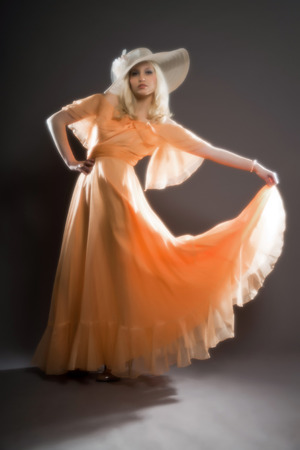Soft focus retro hippie 70s fashion sensual girl with long blonde hair wearing an orange dress and hat. Studio shot against grey.の写真素材