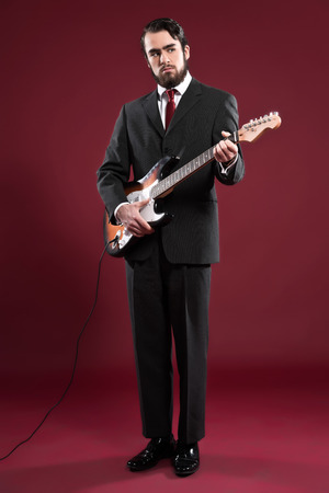 Retro fashion music man with beard wearing grey suit and red tie. Playing electric guitar. Studio shot.の写真素材