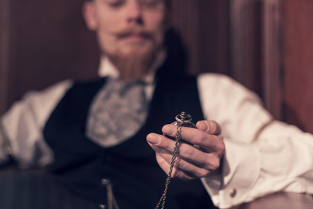 Vintage 1900 fashion man with beard and glasses. Sitting in old wooden reading room. Looking at pocket watch.の写真素材