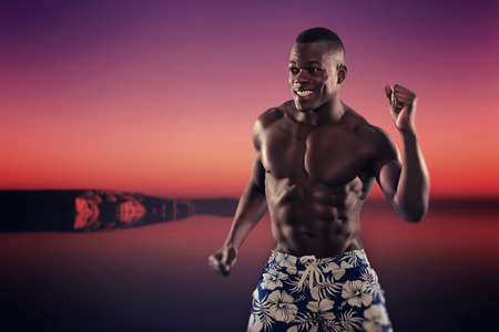 Summer black african american muscled fitness man dancing. Wearing blue swimming shorts. Sunset outdoors at the beach.の写真素材