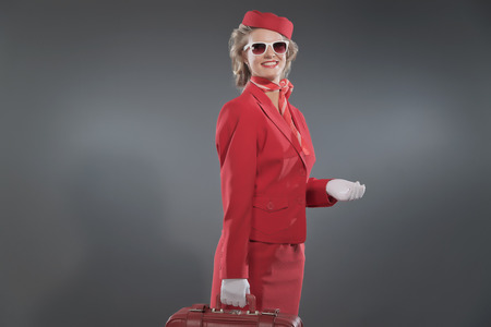 Retro blonde stewardess wearing red suit with cap and sunglasses. Holding red suitcase. Studio shot against grey.の写真素材