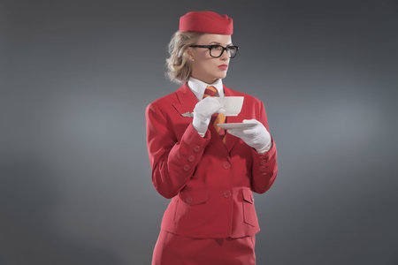 Retro blonde stewardess wearing red suit with striped tie and glasses. Drinking coffee. Studio shot against grey.の写真素材