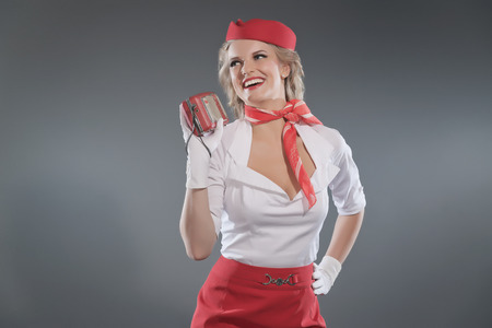 Smiling retro blonde stewardess wearing red skirt and cap with white shirt. Holding vintage radio. Studio shot against grey.の写真素材