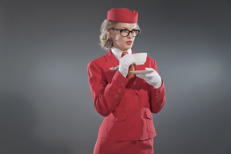Retro blonde stewardess wearing red suit with striped tie and glasses. Drinking coffee. Studio shot against grey.の写真素材