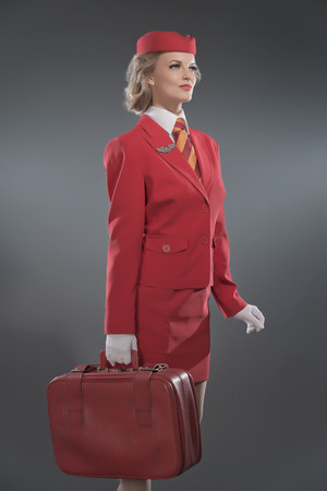 Smiling retro blonde stewardess wearing red suit with striped tie and cap. Holding red suitcase. Studio shot against grey.の写真素材