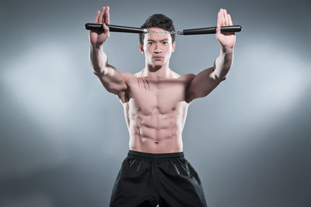 Muscled asian kung fu man in action pose with nunchucks. Blood stripes on his chest. Wearing black pants. Studio shot against grey. の写真素材