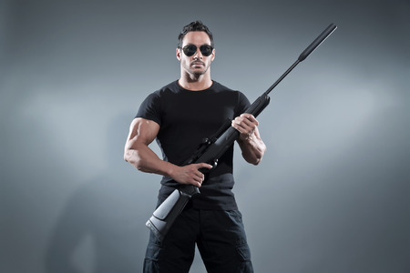 Action hero muscled man holding a rifle. Wearing black t-shirt with pants and sunglasses. Studio shot against grey.の写真素材