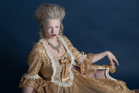Retro baroque fashion woman wearing gold dress. Sitting on vintage couch. Studio shot against blue.