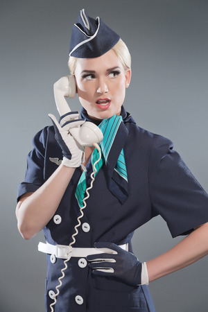 Calling retro blonde stewardess wearing blue suit. Holding white vintage phone. Studio shot against grey.の写真素材