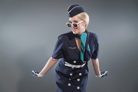 Smiling retro blonde stewardess wearing blue suit. Wearing white sunglasses. Studio shot against grey.の写真素材