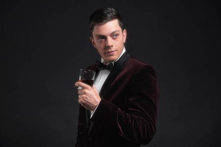 Well dressed good looking business man holding glass of whiskey. Wearing dark red dinner jacket and black bow tie.の写真素材