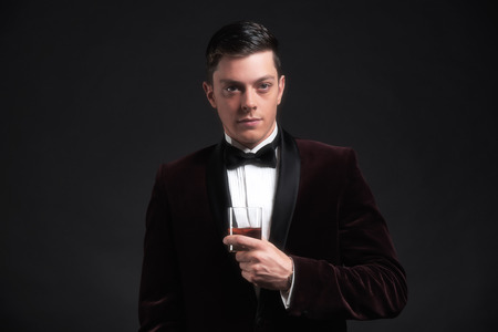 Well dressed good looking business man holding glass of whiskey. Wearing dark red dinner jacket and black bow tie.の写真素材